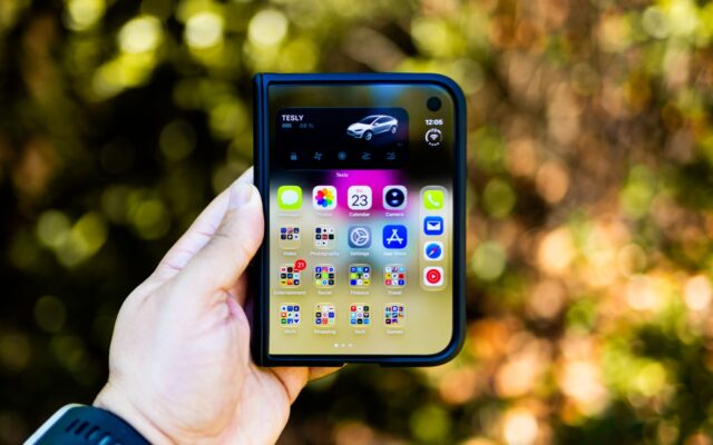Close-up of a hand holding a foldable smartphone showing apps, outdoors with blurred background.
