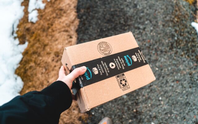 A person holding a cardboard Amazon Prime package on a snowy urban sidewalk.