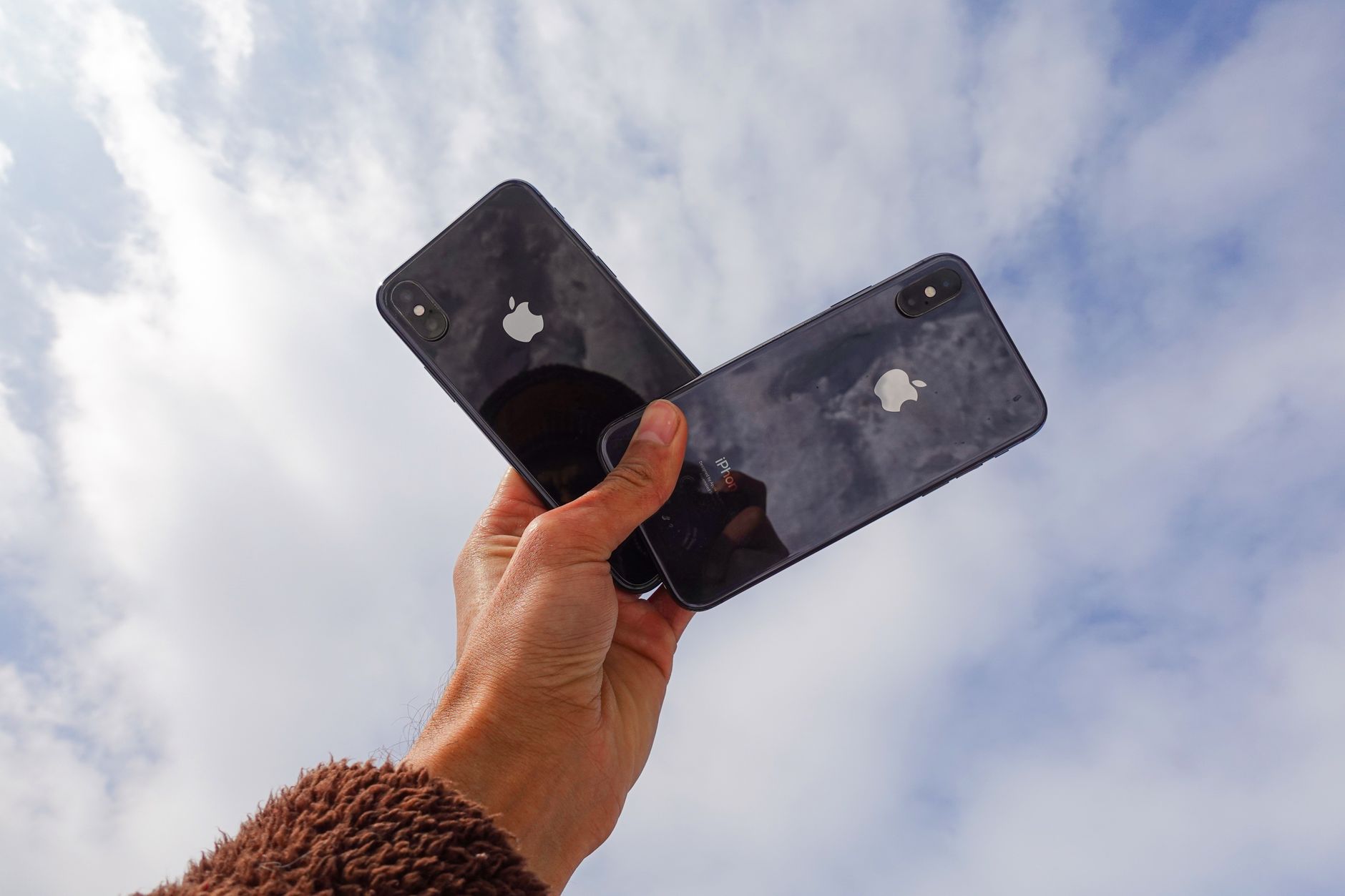 A hand holding two iPhones against a cloudy sky