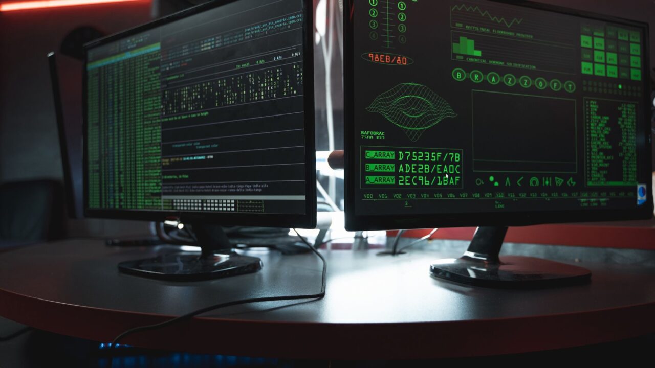 Close-up of dual computer monitors with green coding interfaces in a dark room, highlighting cyber security themes.