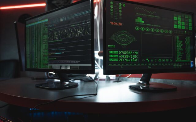 Close-up of dual computer monitors with green coding interfaces in a dark room, highlighting cyber security themes.