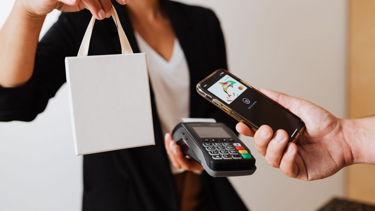 A person uses a smartphone for contactless payment with a payment terminal, while receiving a shopping bag.
