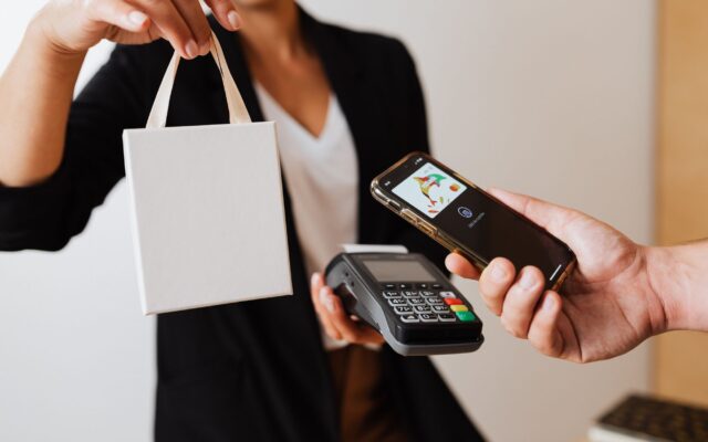 A person uses a smartphone for contactless payment with a payment terminal, while receiving a shopping bag.