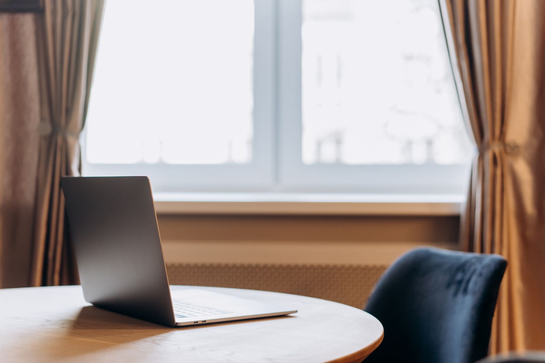 Modern laptop sits on a wooden table with curtains and window in the background.